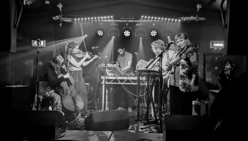 Live performance by a six-piece band with cello, violin, guitar, and electronic equipment, under dramatic stage lighting in a dark venue.